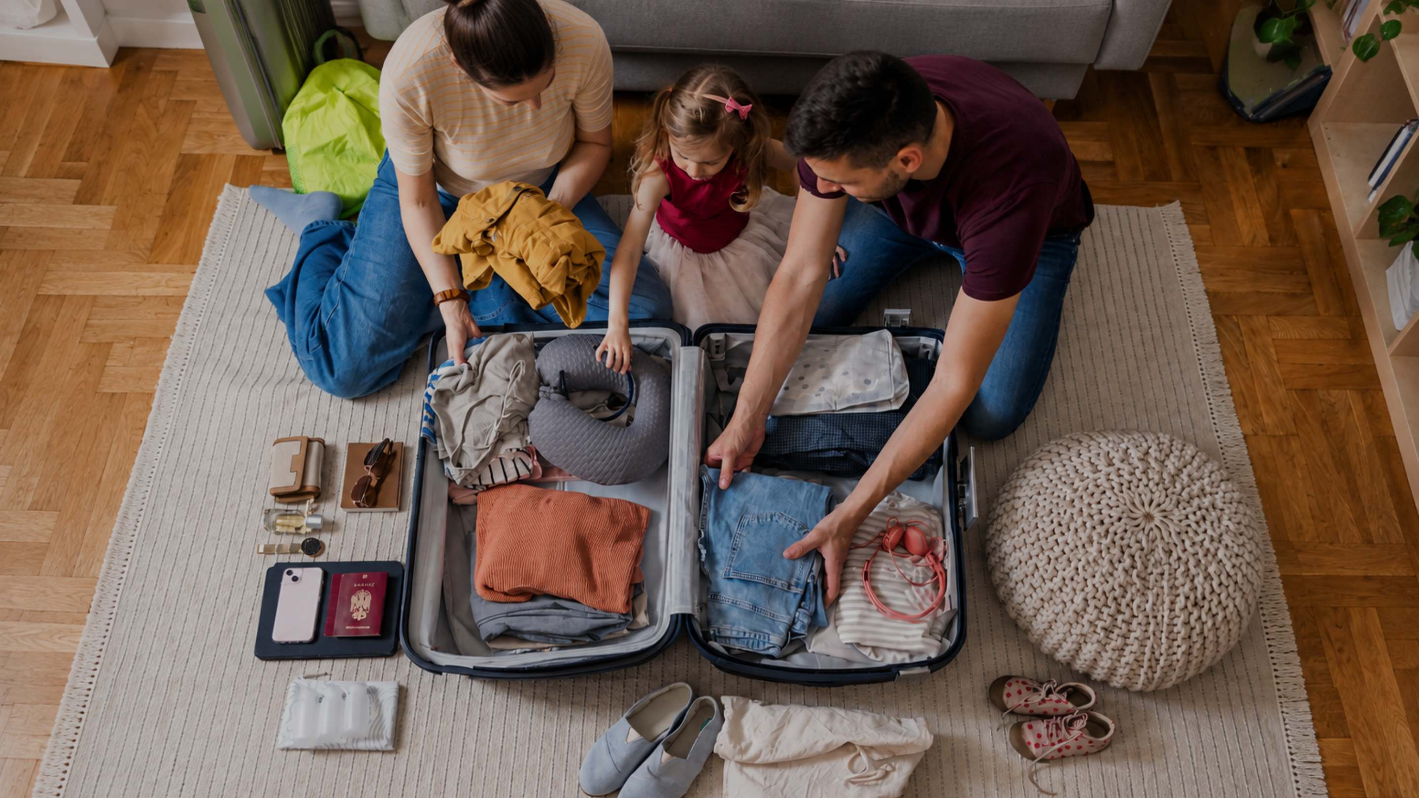 A family of three packing clothes into an open suitcase on a rug in a living room. Folded garments, passports, a phone, and other travel items are arranged on the floor around the suitcase.
