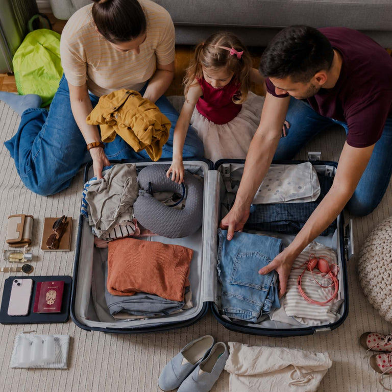A family of three packing clothes into an open suitcase on a rug in a living room. Folded garments, passports, a phone, and other travel items are arranged on the floor around the suitcase.