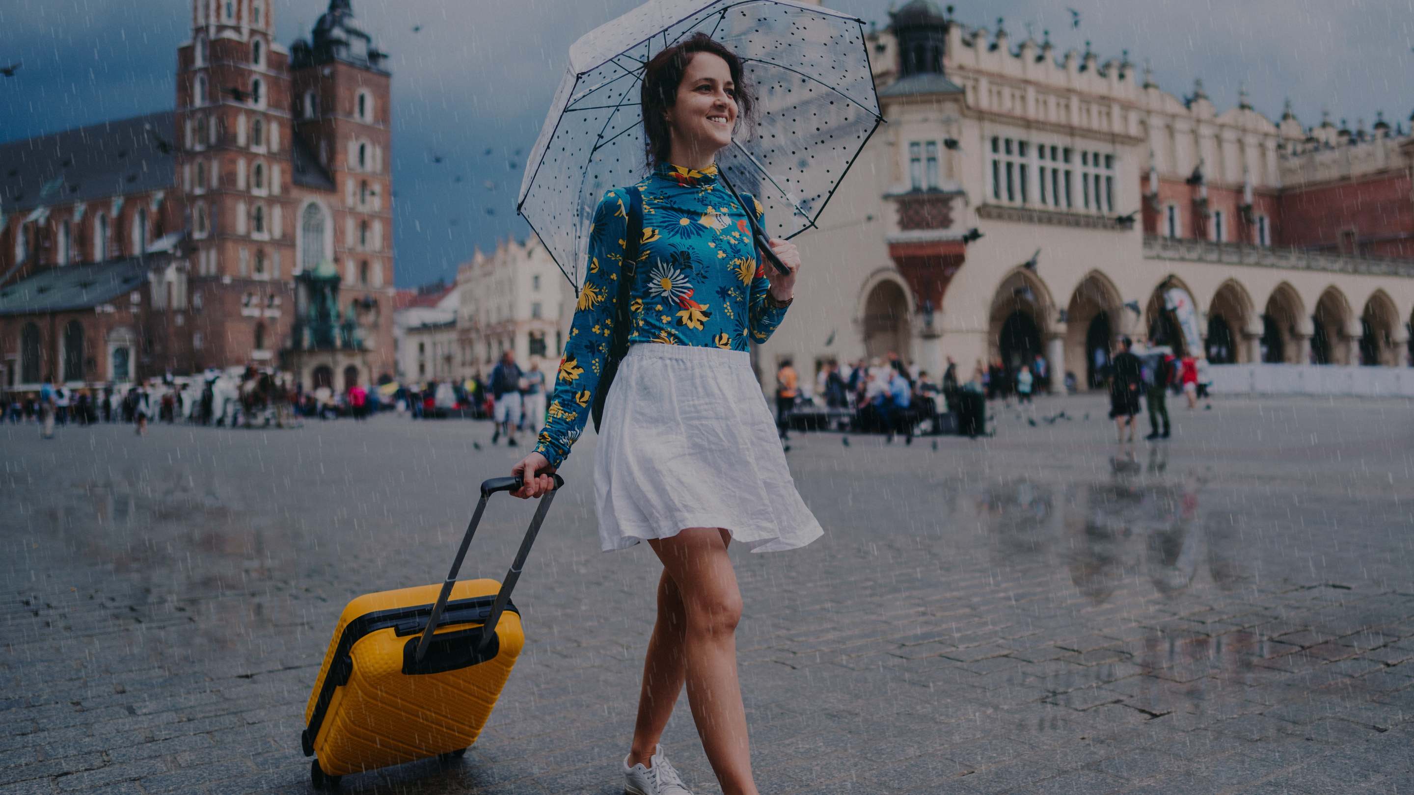 A woman walking in a rainy city square holding a clear umbrella and pulling a yellow rolling suitcase. She is wearing a blue floral long-sleeve top and a white skirt, with historic buildings in the background.